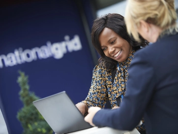 Two female colleagues discussing work together on a grey laptop device.