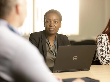Female colleague sitting in a business meeting, listening. A black laptop is in front of the colleague and she does not appear to be saying anything.