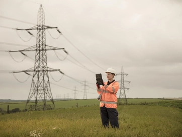 Operative, holding a tablet device up to the air, whilst wearing an orange hi vis jacket in the middle of a field surrounded by electricity pylons.