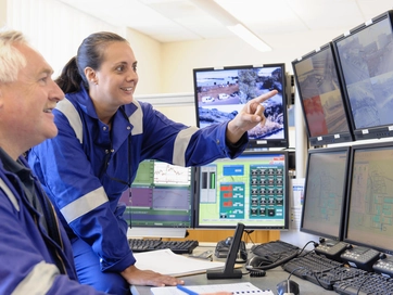 Two colleagues looking at multiple computer screens and spotting information whilst smiling and looking happy. They are both wearing blue overalls.