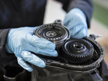 Person handling complex machinery whilst wearing blue gives. The machinery is coated in a black oil substance and is connected via a cog system.