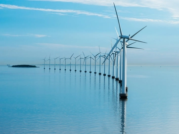 Offshore wind turbines lined up in-front of one another with a blue sky in the distance.