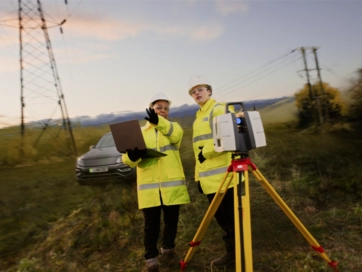 Two women in hi-vis jackets standing in a field and using technical equipment.
