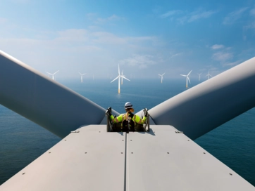 Man standing on top of a wind turbine offshore and out at sea with blue sunny skies.