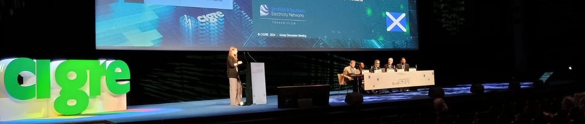 Female attending a conference and speaking at the front of the stage, the room has low lighting and a large suspended presentation slide at the front.