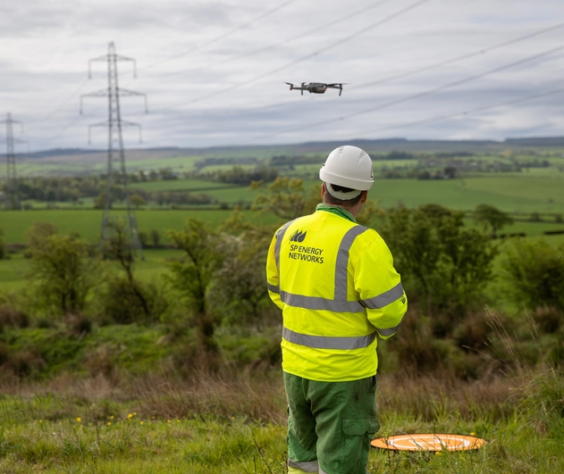 Operative wearing a yellow hi vis jacket flying a drone over a field containing pylons.